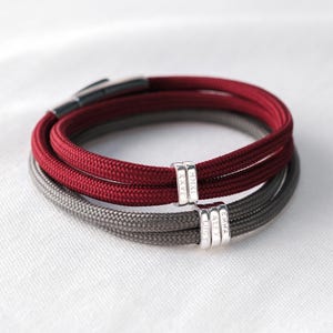 May include: Two bracelets stacked on top of each other. One is burgundy, the other is grey. Each bracelet has a silver band with engraved names. The bracelets are on a white surface.