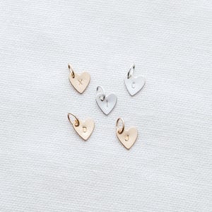 May include: Five gold and silver heart-shaped charms with initial and number engravings. The charms are on a white fabric background.