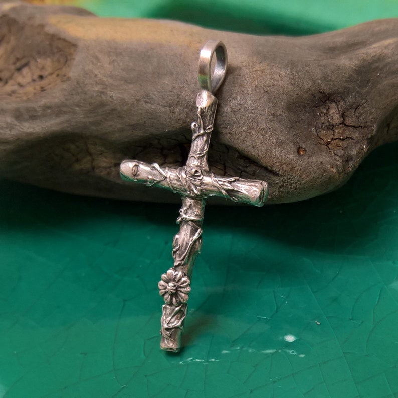 May include: A silver cross pendant with a textured, rustic design. The cross features a floral accent and vine-like details, with a loop at the top for a chain. The pendant is displayed against a natural, dark grey stone.