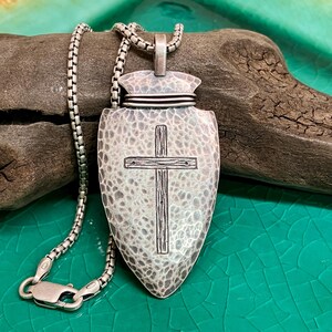 Peut inclure: Un collier pendentif argent&eacute; avec un pendentif en forme de bouclier textur&eacute; et une croix. La cha&icirc;ne est &eacute;galement argent&eacute;e. Le bijou est pr&eacute;sent&eacute; sur du bois flott&eacute;, sur un fond vert. Le pendentif mesure environ 5 cm de long.