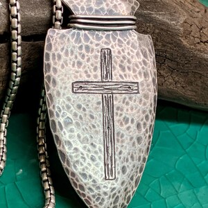 Peut inclure: Un collier pendentif argent&eacute; avec un motif de pointe de fl&egrave;che textur&eacute; et une croix au centre. Le pendentif est attach&eacute; &agrave; une cha&icirc;ne argent&eacute;e. Le fond est un m&eacute;lange de vert et de marron.