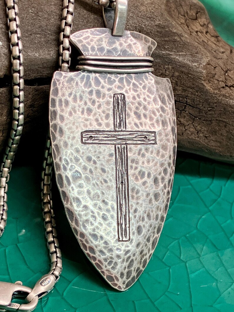 Peut inclure: Un collier pendentif argent&eacute; avec un motif de pointe de fl&egrave;che textur&eacute; et une croix au centre. Le pendentif est attach&eacute; &agrave; une cha&icirc;ne argent&eacute;e. Le fond est un m&eacute;lange de vert et de marron.