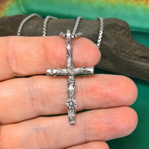 May include: A silver cross pendant with a detailed, textured design. The cross features vine-like patterns and a small flower at the bottom. It hangs from a delicate chain, creating a religious-themed accessory.