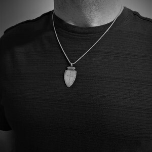Peut inclure: Un collier avec un pendentif argent&eacute; en forme de bouclier orn&eacute; d'une croix. Le pendentif est suspendu &agrave; une fine cha&icirc;ne. Le collier est port&eacute; sur un fond sombre.