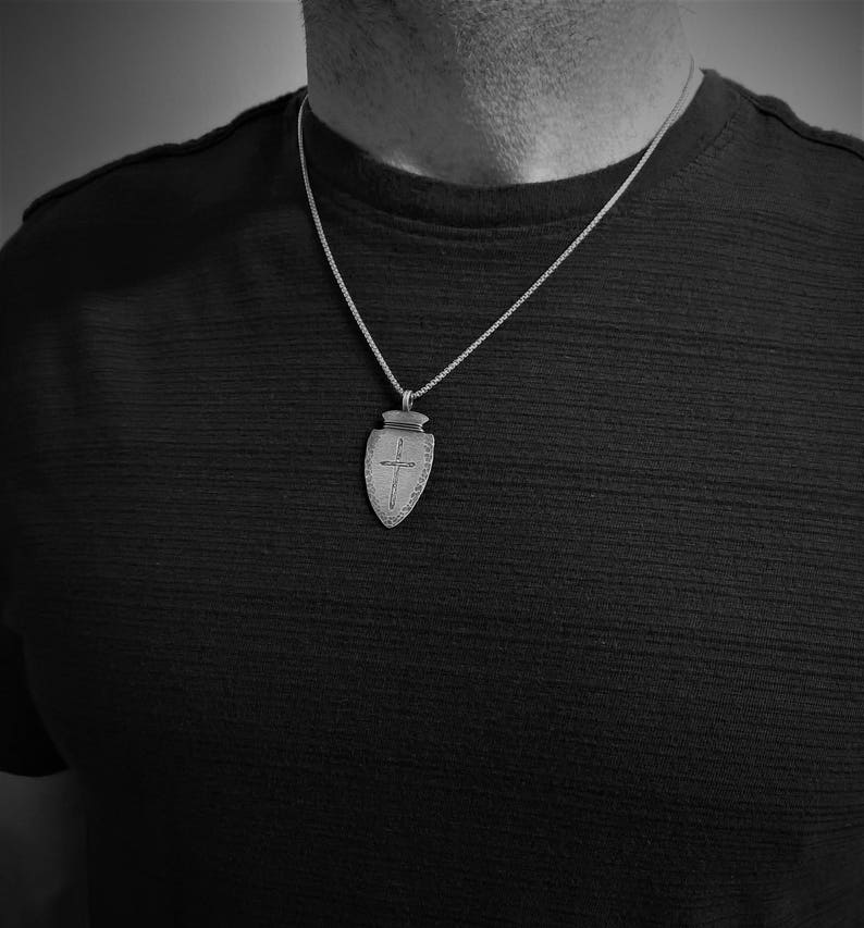 Peut inclure: Un collier avec un pendentif argent&eacute; en forme de bouclier orn&eacute; d'une croix. Le pendentif est suspendu &agrave; une fine cha&icirc;ne. Le collier est port&eacute; sur un fond sombre.