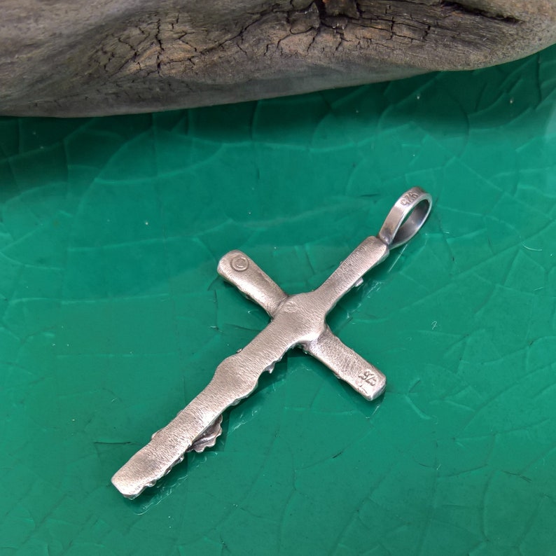 May include: A silver cross pendant with a textured, organic design. The cross has a loop at the top for a chain and is set against a green surface. The pendant is likely made of sterling silver.