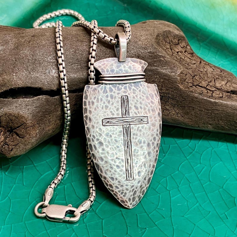 Peut inclure: Un collier avec un pendentif argent&eacute; en forme de pointe de fl&egrave;che textur&eacute;e avec une croix. Le pendentif est suspendu &agrave; une cha&icirc;ne argent&eacute;e. Le collier est pr&eacute;sent&eacute; sur un morceau de bois flott&eacute; sur fond vert.