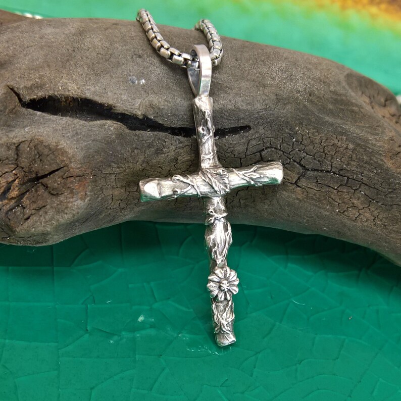 May include: A silver pendant necklace with a detailed cross design. The cross has a textured, rustic appearance with floral and vine-like embellishments. The necklace chain is a linked style.