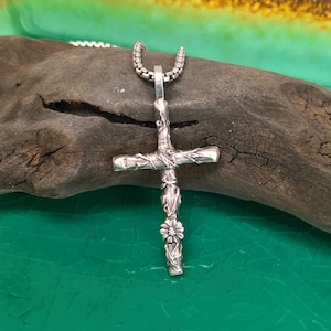 May include: A silver cross pendant with a floral design at the base. The cross features a detailed vine-like pattern and hangs from a silver chain. The pendant is shown against a natural wood background.