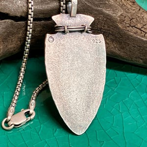 Peut inclure: Un collier avec pendentif argent&eacute; en forme de pointe de fl&egrave;che. Le pendentif pr&eacute;sente une surface textur&eacute;e et est attach&eacute; &agrave; une cha&icirc;ne d&eacute;licate. Le fermoir est visible. Le pendentif est marqu&eacute; "925".