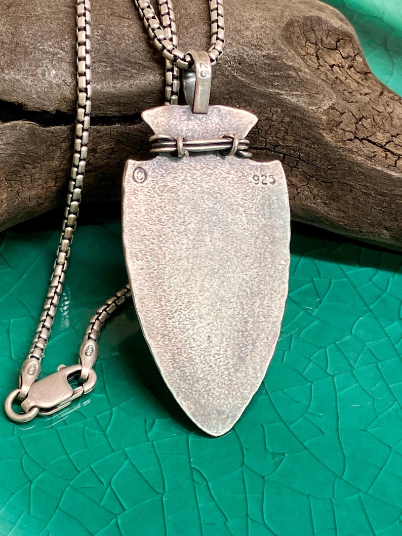 Peut inclure: Un collier avec pendentif argent&eacute; en forme de pointe de fl&egrave;che. Le pendentif pr&eacute;sente une surface textur&eacute;e et est attach&eacute; &agrave; une cha&icirc;ne d&eacute;licate. Le fermoir est visible. Le pendentif est marqu&eacute; "925".