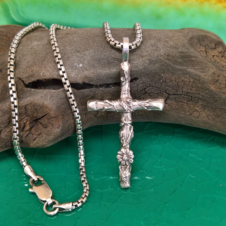 May include: A silver cross pendant necklace with a detailed, textured design. The cross features a floral accent and is suspended from a box chain necklace. The necklace is displayed on a piece of driftwood against a green background.