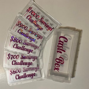 May include: Clear plastic cash box and savings challenge envelopes. The envelopes are labelled with amounts from £600 to £1000, with the text "Savings Challenge" in a pink, glittery font. The cash box has the text "Cash Box" in a similar font.