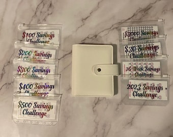 May include: A white planner with a snap closure is surrounded by clear plastic pouches. Each pouch contains a savings challenge card with holographic text. The cards are labeled with amounts like "$100 Savings Challenge" and "2023 Savings Challenge."
