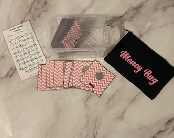 Op de afbeelding: Een doorzichtige plastic container en een zwarte Money Bag met roze tekst worden weergegeven op een marmeren oppervlak. De container bevat verschillende roze en witte zigzagkaarten met een zilveren cirkel. Er is ook een witte kaart met een 52-weekse spaaruitdaging.