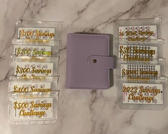 May include: A light purple planner with a snap closure is surrounded by clear plastic pouches with gold lettering. The pouches are labeled with savings challenges, including amounts like $100, $200, $300, $400, and $500.