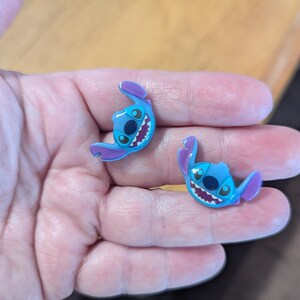 May include: Two blue and purple Stitch character charms. Each charm features Stitch's face with large black eyes, a wide grin, and large ears. The charms are likely for shoes or accessories.