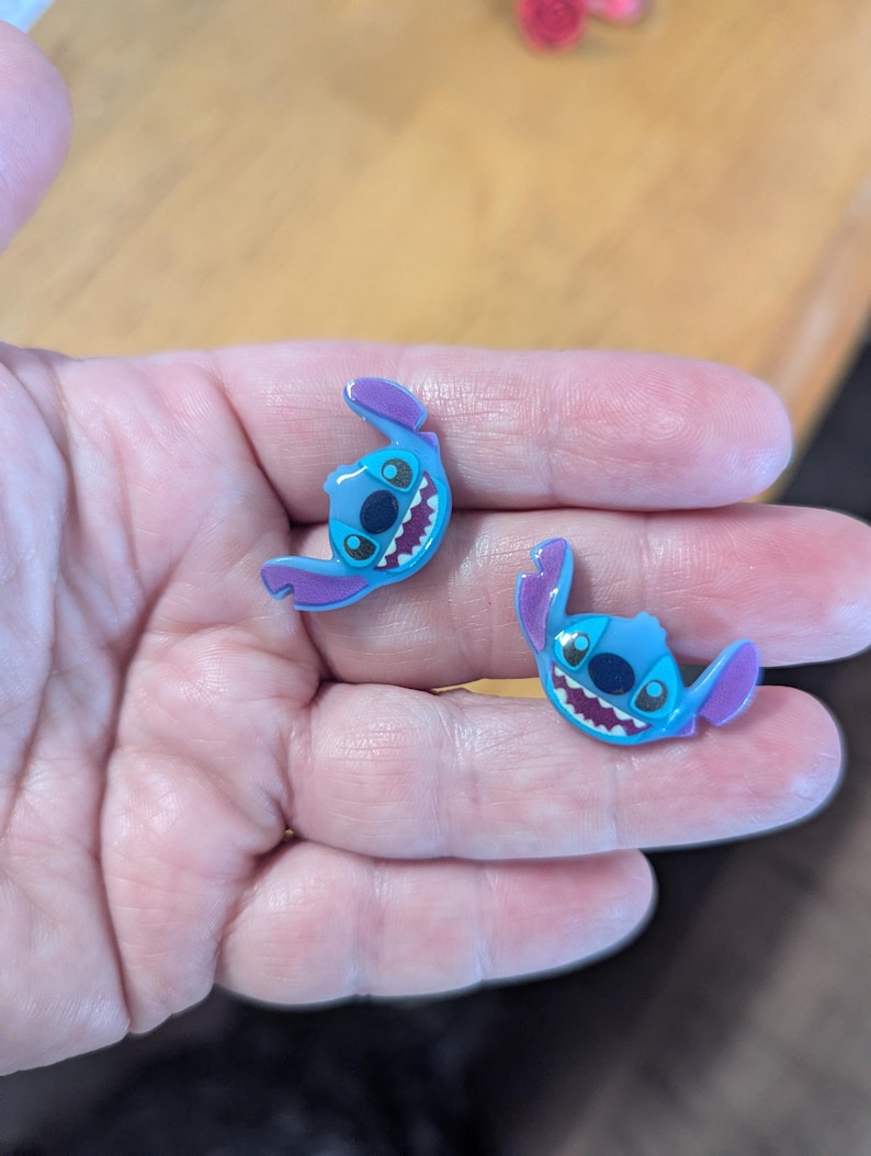 May include: Two blue and purple Stitch character charms. Each charm features Stitch's face with large black eyes, a wide grin, and large ears. The charms are likely for shoes or accessories.
