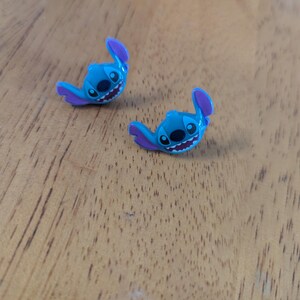 May include: Two small, blue Stitch figurines with purple ears and details, resting on a wooden surface. The figurines have black eyes and a wide, toothy grin, capturing the character's playful expression. These are likely collectible items.
