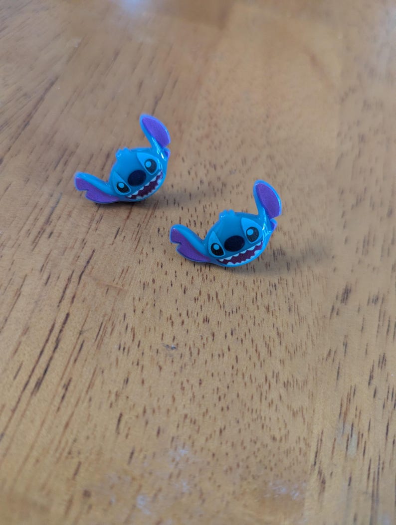 May include: Two small, blue Stitch figurines with purple ears and details, resting on a wooden surface. The figurines have black eyes and a wide, toothy grin, capturing the character's playful expression. These are likely collectible items.