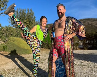 Op de afbeelding: Een stel in kleurrijke kleding. De vrouw draagt een zwarte en witte geprinte legging, een groen topje en een groene franjesjaal. De man draagt een roze en witte geprinte short, een paillettenjas met regenboogkleuren en een gouden ketting.