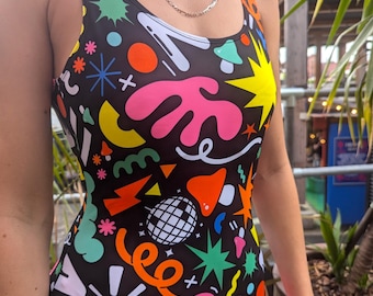 May include: A black one-piece swimsuit with a colorful abstract pattern. The design includes shapes in yellow, pink, orange, green, and white. The swimsuit has a scoop neckline and thin straps.