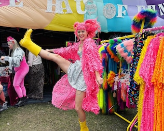 May include: A woman wearing a pink fringed jacket and a silver sequined dress poses with one leg raised, wearing a yellow rain boot. The background features a colorful display of fringed fabric in various colors, including pink, yellow, blue, and green. The sign above the display reads "Haus of Fun".