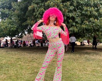 May include: A person wearing a pink and green geometric patterned pantsuit with a pink fuzzy hat and sunglasses. The person is standing in a grassy field.