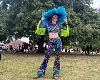 May include: A person wearing a black and neon green patterned outfit with a blue feathered hat and sunglasses. The outfit includes a long-sleeved crop top and flared pants. The person is standing in a grassy field with a large tree in the background.
