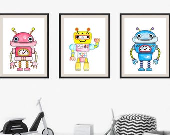 May include: Three framed watercolor robot prints. The robots are in pink, yellow, and blue, each with unique designs and details. The robots have antennas, gauges, and various mechanical features. The prints are displayed on a white wall.