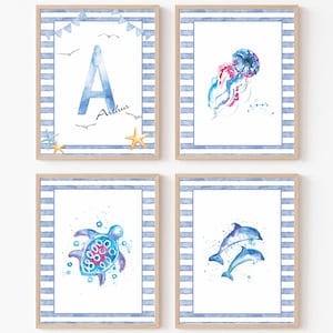 Op de afbeelding: Vier ingelijste wandposters met aquarelillustraties van het onderwaterleven. Eén poster toont de letter A met de naam Arthur, meeuwen en zeesterren. De andere tonen een kwal, een zeeschildpad en twee dolfijnen, allemaal met blauw gestreepte randen.