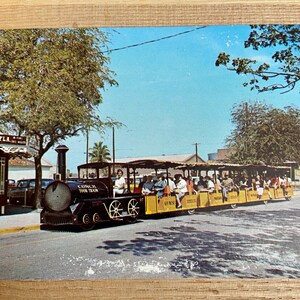 May include: A yellow and black Conch Tour Train with passengers on board. The train is driving down a street with palm trees and buildings in the background.