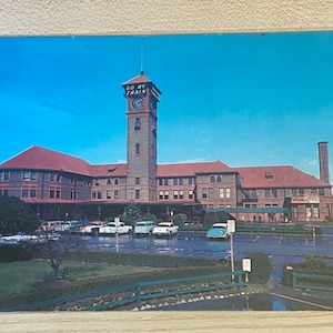 May include: A vintage postcard featuring a photo of a large brick building with a clock tower. The building has a sign that reads "Go By Train". There are several cars parked in front of the building.