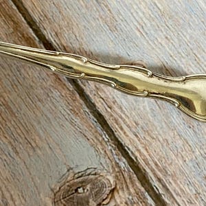 May include: A close-up of a gold-colored serving spoon with ornate detailing. The spoon has a decorative handle with a floral design at the end. The spoon is set against a wooden background.
