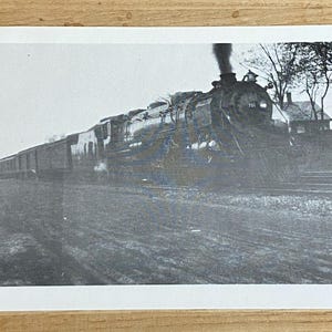 May include: A black and white photograph of a steam locomotive pulling a train of boxcars. The locomotive is emitting smoke and is in motion. The photograph is taken from a low angle, giving the viewer a good view of the locomotive's details.