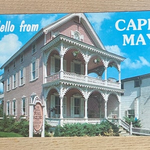 May include: Vintage postcard featuring a pink Victorian house with white trim and a balcony, under a blue sky. Text reads "Hello from CAPE MAY". The postcard also features a sign for "The Pink House Gifts".