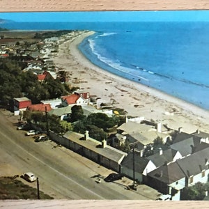 May include: An aerial view of a beach town with white houses and a sandy beach. The ocean is blue and there are waves crashing on the shore. There are trees and a road leading to the beach.
