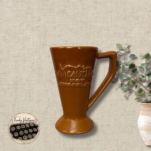 May include: A brown ceramic mug with a flared base and a handle, featuring the embossed text Nesbitt's HOT CHOCOLATE on the front. The mug has a smooth, glossy finish and a tapered shape that widens toward the top rim.