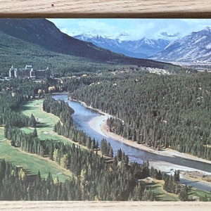 May include: Vintage postcard featuring a scenic aerial view of a river winding through a lush green forest. Snow-capped mountains rise in the background, with a large building nestled among the trees. The postcard has a retro aesthetic.