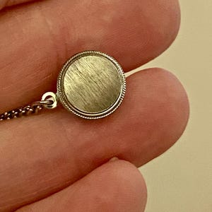 May include: A small, round, silver-toned pendant with a textured surface and a beaded edge. The pendant is attached to a delicate chain. The image is a close-up, highlighting the pendant's details.