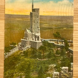 May include: A postcard depicting the Will Rogers Shrine of the Sun on Cheyenne Mountain in Colorado Springs, Colorado. The shrine is a stone tower with a pointed roof and is surrounded by a stone wall and greenery.