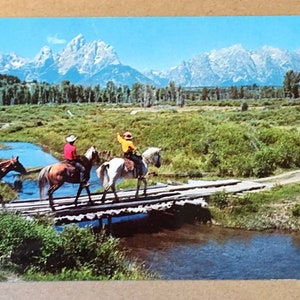 May include: Three people on horseback ride across a wooden bridge over a stream. The horses are brown and white. The people are wearing cowboy hats and western clothing. The background features a mountain range with snow-capped peaks.