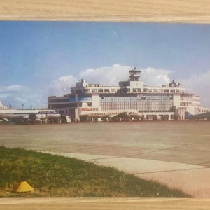 May include: Vintage postcard featuring an airport terminal building with a control tower, airplanes, and a blue sky with clouds. The terminal has a modern design with large windows. The postcard is on a wooden surface.