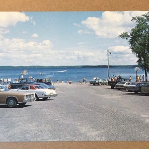 May include: A vintage postcard showing a beach scene with a lake, a sandy shore, and a parking lot filled with cars. People are swimming and sunbathing on the beach. The sky is blue with white clouds.