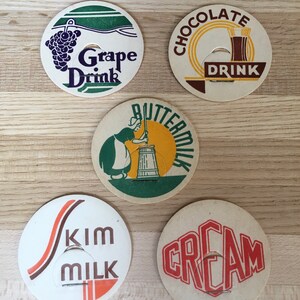 May include: A collection of vintage paper drink coasters. The coasters feature graphics and text for Grape Drink, Chocolate Drink, Buttermilk, Skim Milk, and Cream. Each coaster has a unique design with a central cutout.
