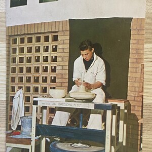 May include: A young person is working on a pottery wheel, shaping a clay pot. The wheel is on a wooden stand with a shelf underneath. There is a white towel and a bucket on the shelf. The person is wearing a white shirt and white pants. The background is a brick wall with a window.