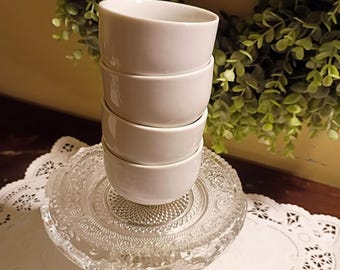 May include: A stack of four plain white ceramic bowls resting on a clear glass pedestal dish. The bowls are smooth and cylindrical with a simple rim. The glass pedestal features a textured, pressed pattern on its surface and a sturdy base.