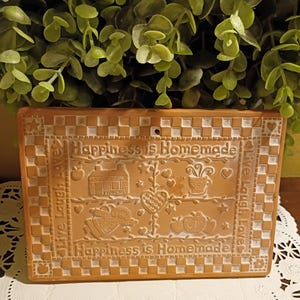 May include: A rectangular terracotta plaque featuring a checkered border and the embossed text "Happiness is Homemade" and "Live Laugh Love." The center displays carved icons including a house, a heart, a cup, an angel, and apples.