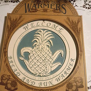 May include: A square bread and bun warmer in tan cardboard packaging. The center features a circular teal and cream pineapple graphic with the text WELCOME, THE SYMBOL OF HOSPITALITY, and BREAD AND BUN WARMER. Wheat stalks and garlic bulbs decorate the corners.
