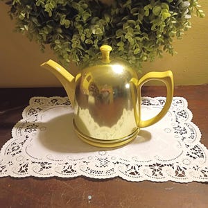 May include: A shiny, gold-colored teapot with yellow accents, including the handle, spout, and lid. The teapot sits on a white, lacy doily. A green wreath is in the background.